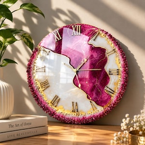 Può includere: Un orologio da parete rotondo con una superficie lucida divisa tra magenta intenso e bianco, accentuata da venature dorate. L'orologio presenta numeri romani dorati e lancette sottili dorate. Il bordo esterno è texturizzato con piccoli cristalli rosa scuro.