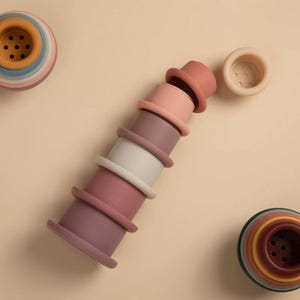 Puede incluir: Un juego de vasos apilables de colores en tonos rosa, blanco y amarillo. Los vasos están dispuestos en una torre, con algunas piezas dispersas. El juguete está diseñado para bebés y niños pequeños.