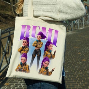 May include: A beige tote bag featuring the name "RUMI" in purple block letters. The bag displays a cartoon illustration of a woman with purple hair in a yellow jacket and black pants. The woman is shown in multiple poses.