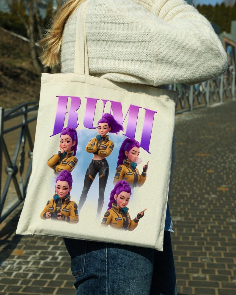 May include: A beige tote bag featuring the name "RUMI" in purple block letters. The bag displays a cartoon illustration of a woman with purple hair in a yellow jacket and black pants. The woman is shown in multiple poses.