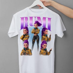 May include: White t-shirt featuring the name "RUMI" in purple and a graphic of a character with purple hair and a yellow jacket. The character is shown in multiple poses. The shirt is hanging on a white hanger.
