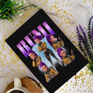 May include: Black notebook featuring the name "RUMI" in large purple letters. The notebook's design includes multiple images of a woman with purple hair wearing a yellow jacket and black pants. The background is a light blue sky.