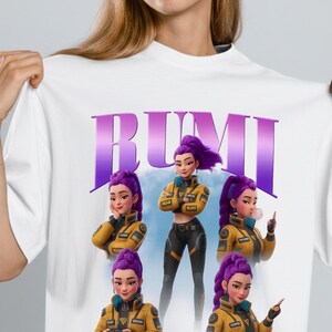 May include: White t-shirt featuring the word "RUMI" in purple block letters above a graphic of a woman with purple hair in a yellow jacket and black pants. The graphic includes multiple poses of the woman.