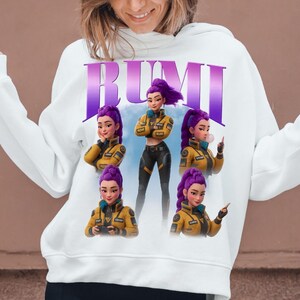 May include: White hoodie featuring the word "RUMI" in purple letters. The graphic design includes a cartoon character with purple hair in a yellow jacket and black pants. The character is shown in multiple poses.