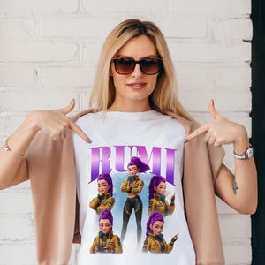 May include: White t-shirt featuring the word "RUMI" in purple, with illustrations of a woman with purple hair and a yellow jacket. The woman in the image is wearing sunglasses and pointing at the shirt.