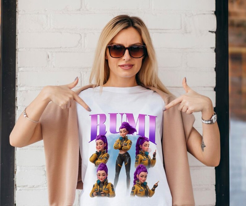 May include: White t-shirt featuring the word "RUMI" in purple, with illustrations of a woman with purple hair and a yellow jacket. The woman in the image is wearing sunglasses and pointing at the shirt.