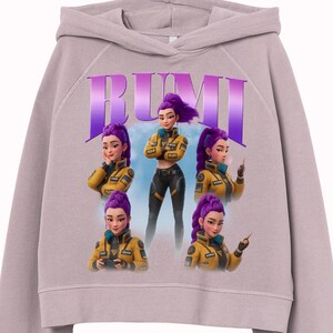 May include: A light purple cropped hoodie featuring a graphic of a woman with purple hair in a yellow jacket. The word "RUMI" is printed in large purple letters above the graphic. The graphic shows the woman in different poses.