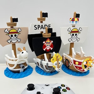 Może przedstawiać: Trzy zabawki w postaci statków pirackich z różnymi wzorami i kolorami żagli, w tym czaszką i skrzyżowanymi piszczelami, płonącą czaszką i napisem "SPADE". Każdy statek stoi na niebieskiej podstawie w kształcie fali. Biały kontroler Xbox znajduje się na pierwszym planie.