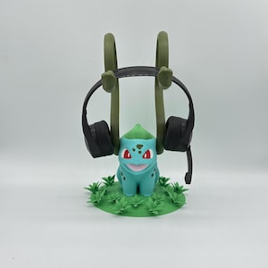 May include: A teal Bulbasaur-themed headphone stand with a green base and accents. The stand holds a pair of black headphones. The Bulbasaur figure has red eyes and a smiling mouth. The stand is designed to hold headphones.