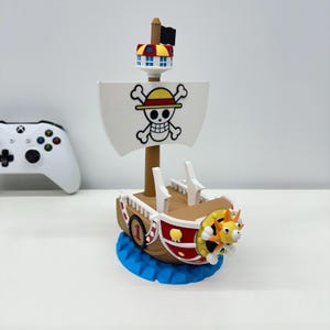 Może przedstawiać: Szczegółowy model statku z białym żaglem z emblematem czaszki i skrzyżowanych piszczeli. Statek ma brązowy kadłub, czerwone akcenty i żółtą figurę dziobową. Stoi na niebieskiej podstawie, z małą wieżą i czarną flagą.