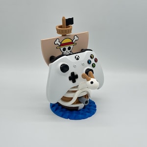 Może przedstawiać: Biały kontroler Xbox spoczywa na dekoracyjnym stojaku w kształcie statku. Stojak posiada beżowy żagiel z logo czaszki, brązowy maszt i czarną flagę. Podstawa jest niebieska.