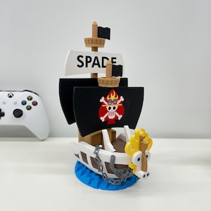 Może przedstawiać: Drewniany model statku pirackiego z czarnymi żaglami i emblematem czaszki i skrzyżowanych piszczeli. Statek ma napis "SPADE" na białym znaku. Statek stoi na niebieskiej podstawie i ma żółtą figurę dziobową smoka.