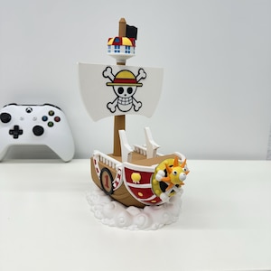 Może przedstawiać: Szczegółowa figurka statku z białym żaglem z motywem czaszki i skrzyżowanych piszczeli. Statek ma brązowy kadłub, czerwone akcenty oraz żółto-pomarańczową figurę dziobową. Statek jest zamontowany na podstawie w kształcie chmury.