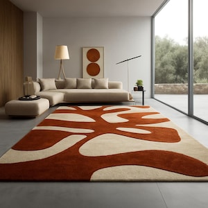 May include: A large, rectangular area rug with a modern design. The rug features a rust-colored, organic pattern against a cream-colored background. The rug is placed in a living room setting with a beige sofa and large windows.