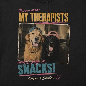 May include: A black t-shirt featuring a graphic of a golden retriever and a black Labrador, both wearing baseball caps. The text reads: These are MY THERAPISTS and I pay in SNACKS! Cooper & Shadow with a small pink heart icon below the text.