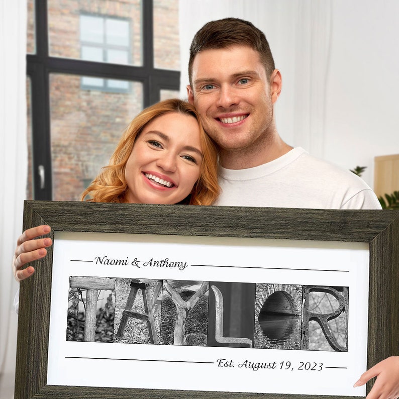 May include: A couple holding a framed black and white photo print. The print spells out with different alphabet photography images the last name 'TAYLOR'. The text above the name reads 'Naomi & Anthony.' The text below the name reads 'Est. August 19, 2023.'