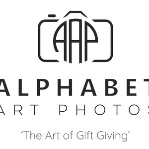 May include: Black and white logo for Alphabet Art Photos, a photography business. The logo features a stylized camera with the letters "AAP" inside. The text "Alphabet Art Photos" is below the camera, and the tagline "The Art of Gift Giving" is below that.