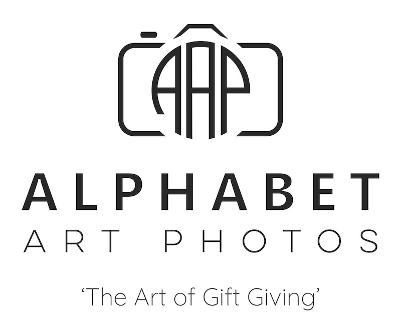 May include: Black and white logo for Alphabet Art Photos, a photography business. The logo features a stylized camera with the letters "AAP" inside. The text "Alphabet Art Photos" is below the camera, and the tagline "The Art of Gift Giving" is below that.