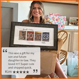 May include: A framed print with a personalized design featuring the names "Garrett & Lindsay" and the dates "EST 10 20 23" with the word "Miller" below. The print is in a dark wood frame and is being held by a person.