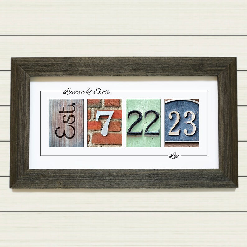May include: A framed print with the text "Lauren & Scott - Est. 7 22 23 - Lee" in a black frame. The print features a collage of four images: a wooden sign with the word "Est.", a brick wall with the number "7", a green wall with the number "22", and a blue sign with the number "23".