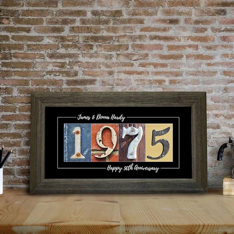 A framed collage style print with colorful photo numbers (1975) taken from buildings, mail boxes, walls etc. The print is personalized with a couple's first and last names, as well as the following sentiment 'Happy 50th Anniversary'. The print has a black background and is framed in a charcoal gray barn wood frame. The frame is sitting on a wooden countertop against a red brick wall.