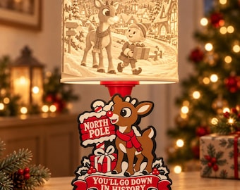 May include: A table lamp featuring a cylindrical shade with a winter scene of a reindeer and an elf. The base is a cutout of a reindeer wearing a red scarf next to a North Pole sign and a gift, with the text "NORTH POLE" and "YOU'LL GO DOWN IN HISTORY".
