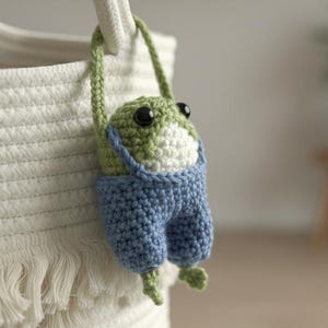 May include: A handmade crochet frog toy with a green and white head, black eyes, and blue overalls. The frog is hanging from a white rope, attached to a white basket with a textured design. The frog's arms and legs are green.