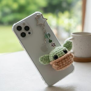 May include: A clear phone case with a green and beige crocheted character charm. The charm is attached to the phone with a silver chain and star-shaped charm. The background is blurred, showing a window and a white surface.
