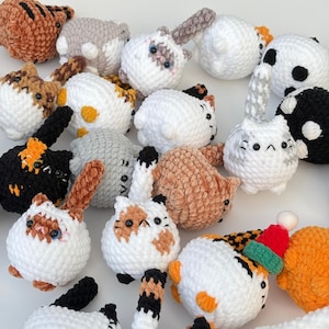 Puede incluir: Una colecci&oacute;n de juguetes de gato hechos a mano con ganchillo en varios colores y patrones. Los juguetes presentan cuerpos blancos, negros, naranjas y grises con rasgos faciales detallados y colas erguidas. Algunos gatos tienen marcas de calic&oacute;, otros son de colores lisos.