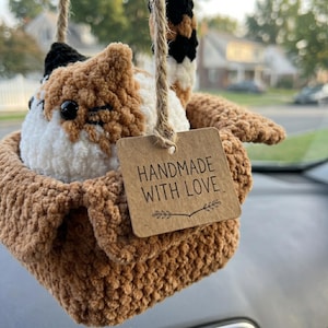 Pu&ograve; includere: Un peluche a forma di gatto calico all'uncinetto seduto dentro una piccola scatola marrone all'uncinetto. Il gatto ha pelo in filato nero, bianco e arancione con occhi in plastica nera. Un cartellino di cartone riporta HANDMADE WITH LOVE.