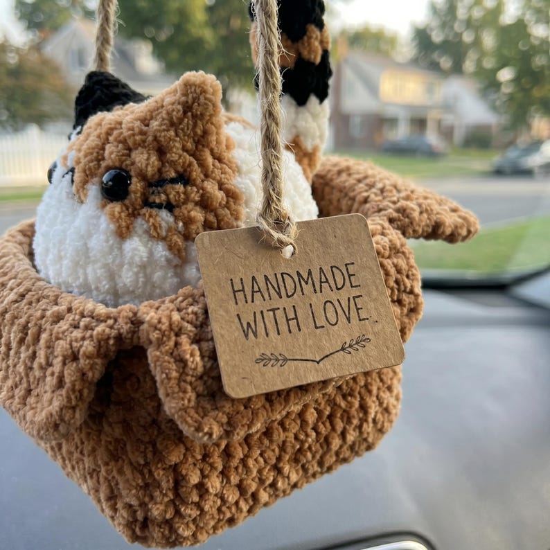 Pu&ograve; includere: Un peluche a forma di gatto calico all'uncinetto seduto dentro una piccola scatola marrone all'uncinetto. Il gatto ha pelo in filato nero, bianco e arancione con occhi in plastica nera. Un cartellino di cartone riporta HANDMADE WITH LOVE.