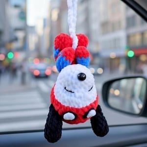 May include: A handmade, crocheted car charm in the shape of a smiling panda. The panda has a white face, red and black accents, and blue and red ears. It hangs from a white cord, perfect for adding a touch of whimsy to any vehicle.