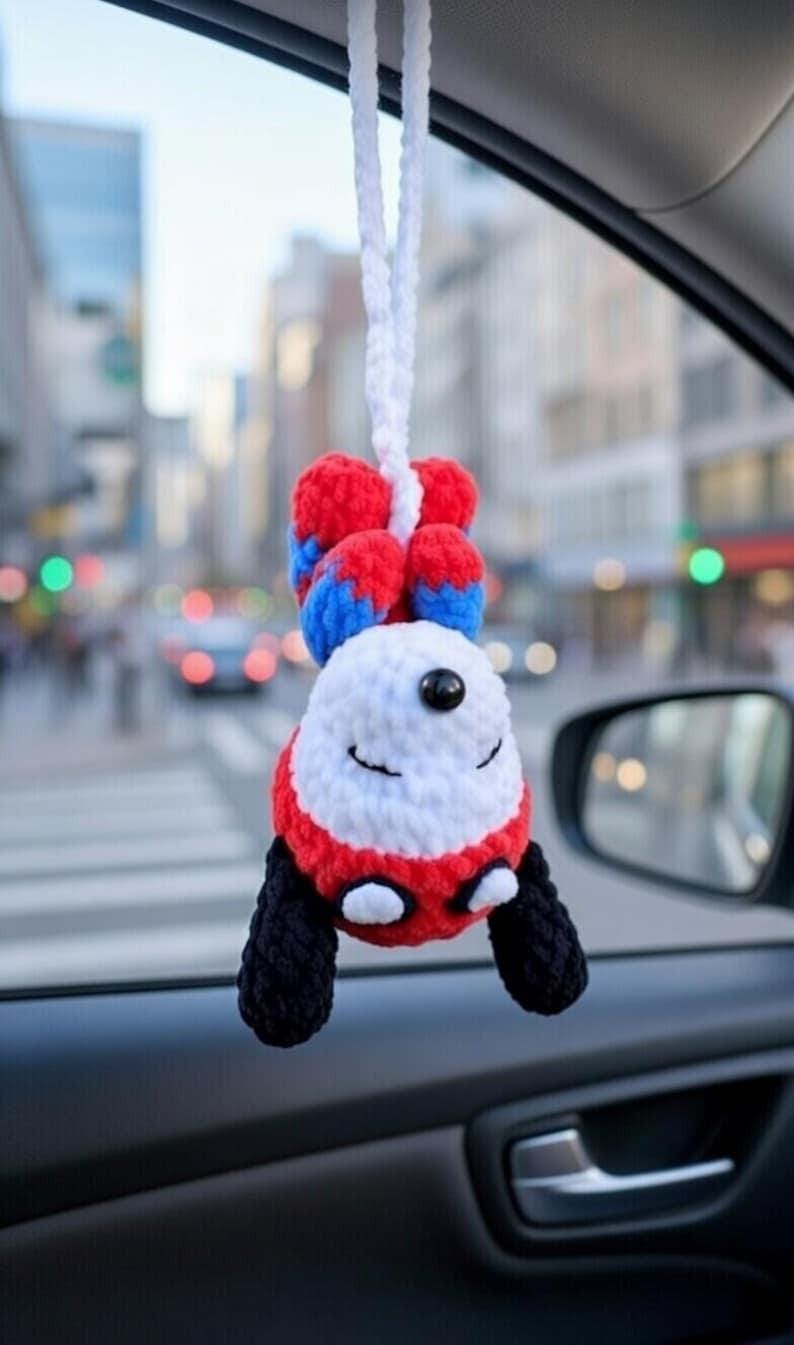 May include: A handmade, crocheted car charm in the shape of a smiling panda. The panda has a white face, red and black accents, and blue and red ears. It hangs from a white cord, perfect for adding a touch of whimsy to any vehicle.