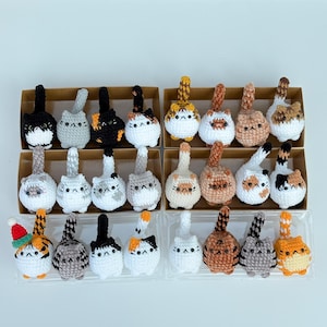 Puede incluir: Una colecci&oacute;n de adornos de gato de ganchillo hechos a mano en varios colores y patrones. Cada gato tiene un dise&ntilde;o &uacute;nico, incluyendo negro, gris, naranja y blanco. Algunos gatos tienen sombreros festivos. Los adornos se exhiben en peque&ntilde;as cajas de cart&oacute;n y recipientes de pl&aacute;stico transparente.