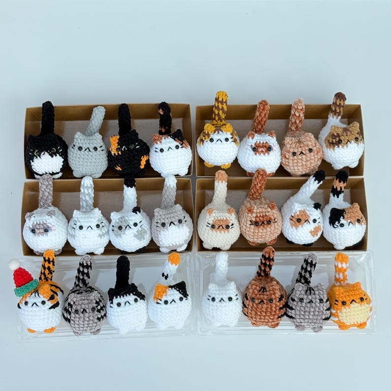Puede incluir: Una colecci&oacute;n de adornos de gato de ganchillo hechos a mano en varios colores y patrones. Cada gato tiene un dise&ntilde;o &uacute;nico, incluyendo negro, gris, naranja y blanco. Algunos gatos tienen sombreros festivos. Los adornos se exhiben en peque&ntilde;as cajas de cart&oacute;n y recipientes de pl&aacute;stico transparente.