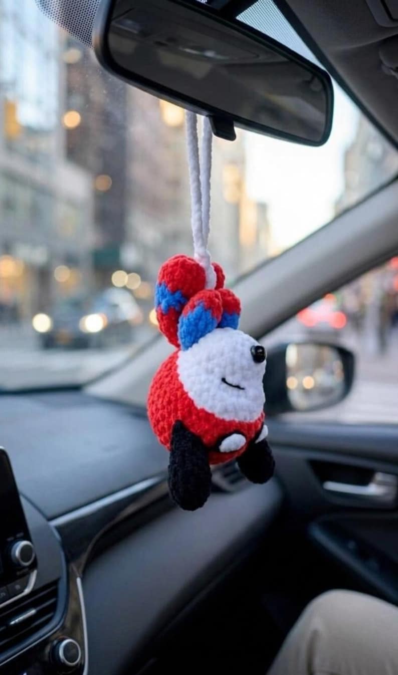 May include: A handmade crochet car charm in the shape of a fox. The fox has a red body, white face, and black paws. The ears are red with blue stripes. It hangs from a white cord.