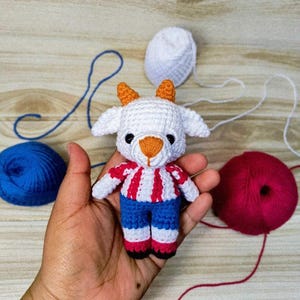 May include: A small crocheted goat figurine featuring a white head with orange horns and nose, black eyes, and a body wearing a red and white striped shirt with blue pants. The toy is crafted with yarn and displays detailed stitching throughout its form.