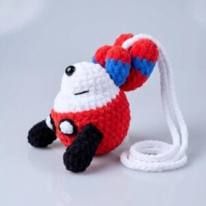 May include: A handmade crochet plush toy in the shape of a cartoon character. The toy features a white head, red body, black feet, and red and blue accents on the ears. A white cord is attached to the toy.