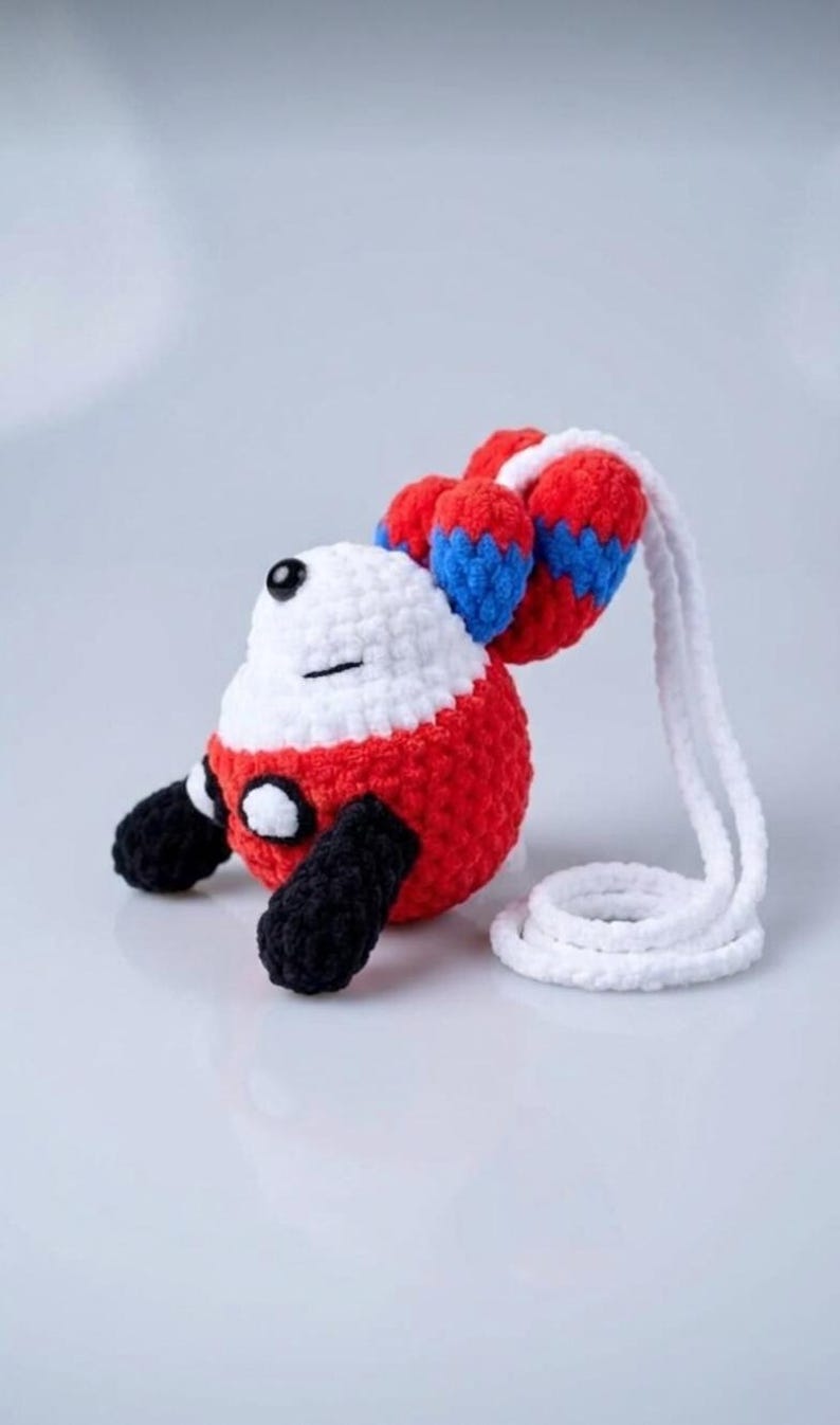 May include: A handmade crochet plush toy in the shape of a cartoon character. The toy features a white head, red body, black feet, and red and blue accents on the ears. A white cord is attached to the toy.