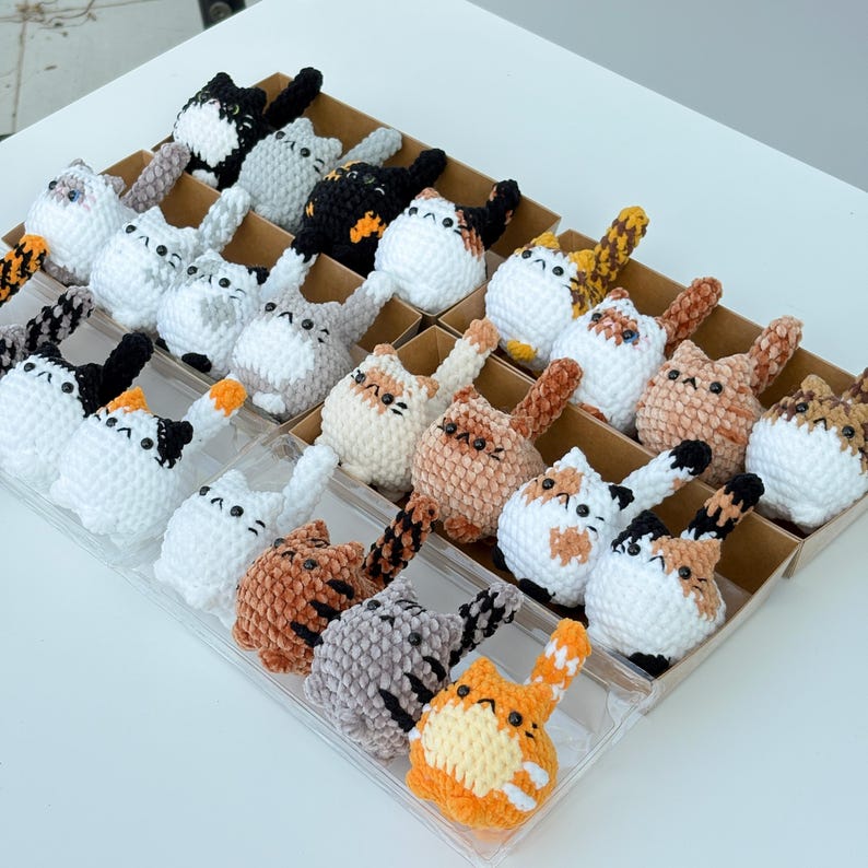 Puede incluir: Una colecci&oacute;n de figuras de gato de ganchillo hechas a mano en varios colores, incluyendo blanco, negro, naranja y gris. Cada gato tiene un patr&oacute;n &uacute;nico y est&aacute; dispuesto en compartimentos individuales dentro de una bandeja.