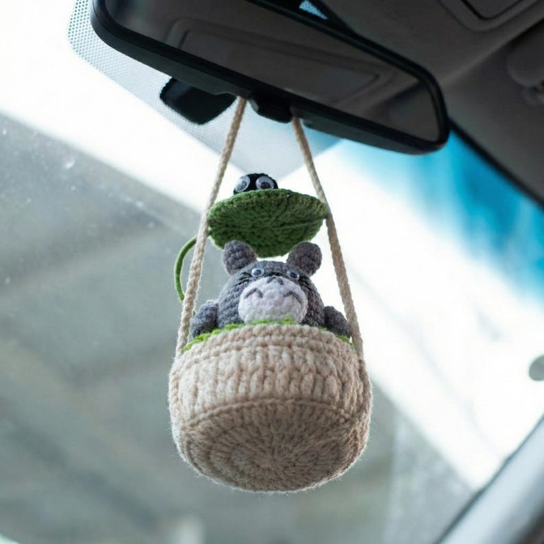 Może przedstawiać: Ręcznie robiony szydełkowy urok do samochodu z szarą postacią Totoro w beżowym koszu. Zielony liść i ruchome oczy znajdują się nad koszem. Urok wisi na lusterku wstecznym samochodu.