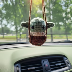 May include: A handmade crocheted figure of a baby character in a swing, in shades of green and brown. The car accessory is suspended from a brown string, hanging in front of a car dashboard. The item is inspired by a popular sci-fi franchise.