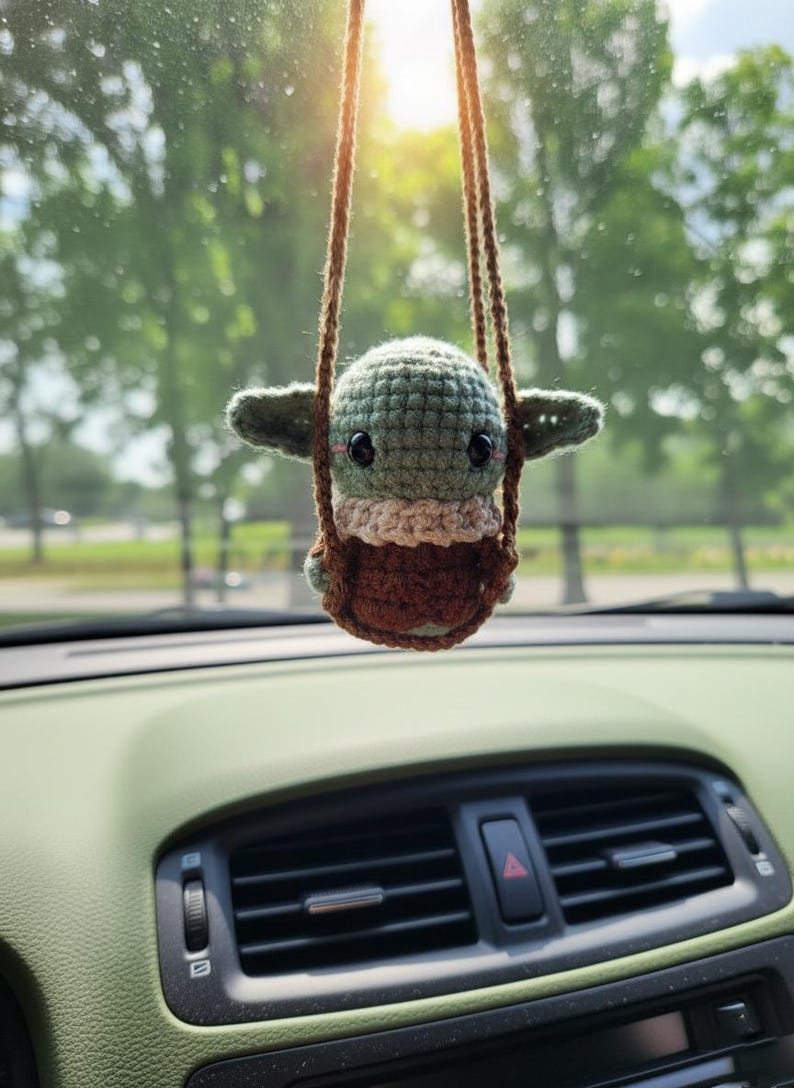 May include: A handmade crocheted figure of a baby character in a swing, in shades of green and brown. The car accessory is suspended from a brown string, hanging in front of a car dashboard. The item is inspired by a popular sci-fi franchise.