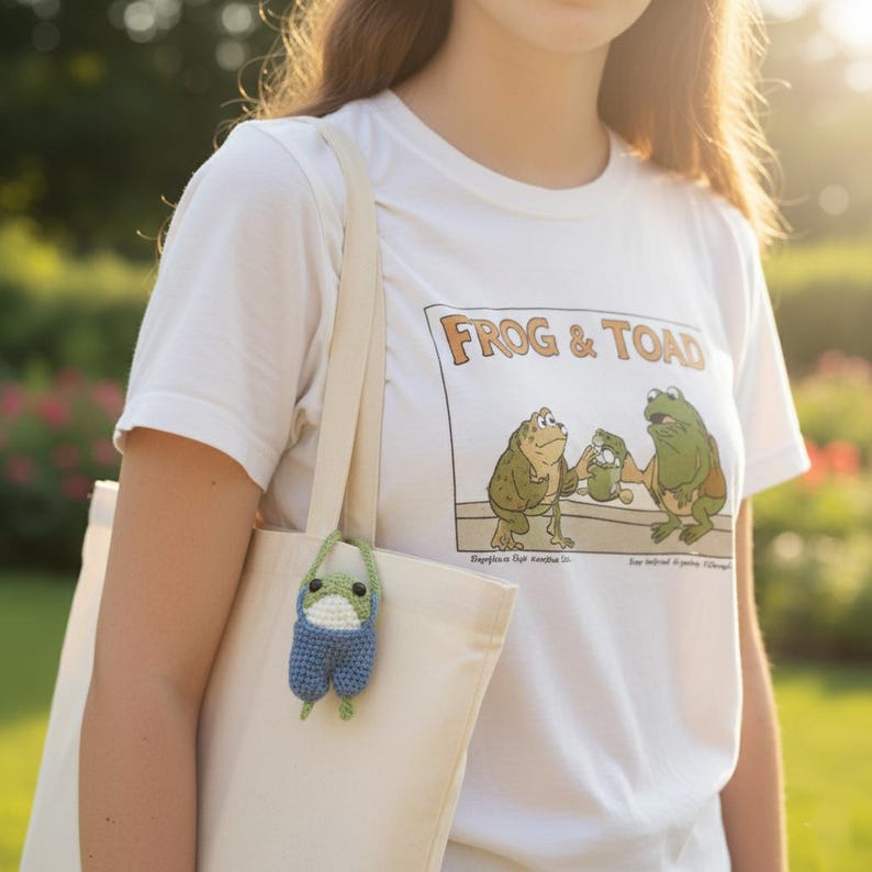 May include: White t-shirt with a "Frog & Toad" graphic, paired with a cream-colored tote bag. A small, blue and white crocheted frog charm hangs from the bag. The shirt features a cartoon illustration of two frogs. The background is a blurred outdoor scene.