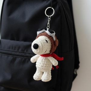 May include: A handmade crochet Snoopy keychain, featuring a white body, black nose, and a red scarf. The keychain has a brown aviator hat with goggles. It is attached to a black backpack.