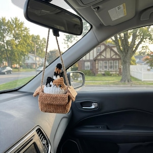 Pu&ograve; includere: Un peluche a forma di gatto calico all'uncinetto seduto dentro una piccola scatola marrone all'uncinetto, appeso a un cordino beige allo specchietto retrovisore di un'auto. Il gatto presenta filati bianchi, arancioni e neri con una coda.