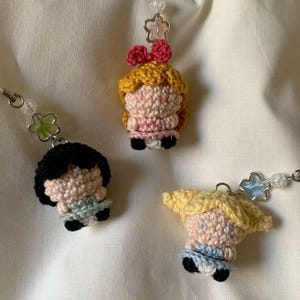 May include: Three handmade crochet doll keychains. Each doll has a unique hairstyle and outfit, with colors including pink, yellow, black, and blue. The keychains feature beaded accents and star-shaped charms.