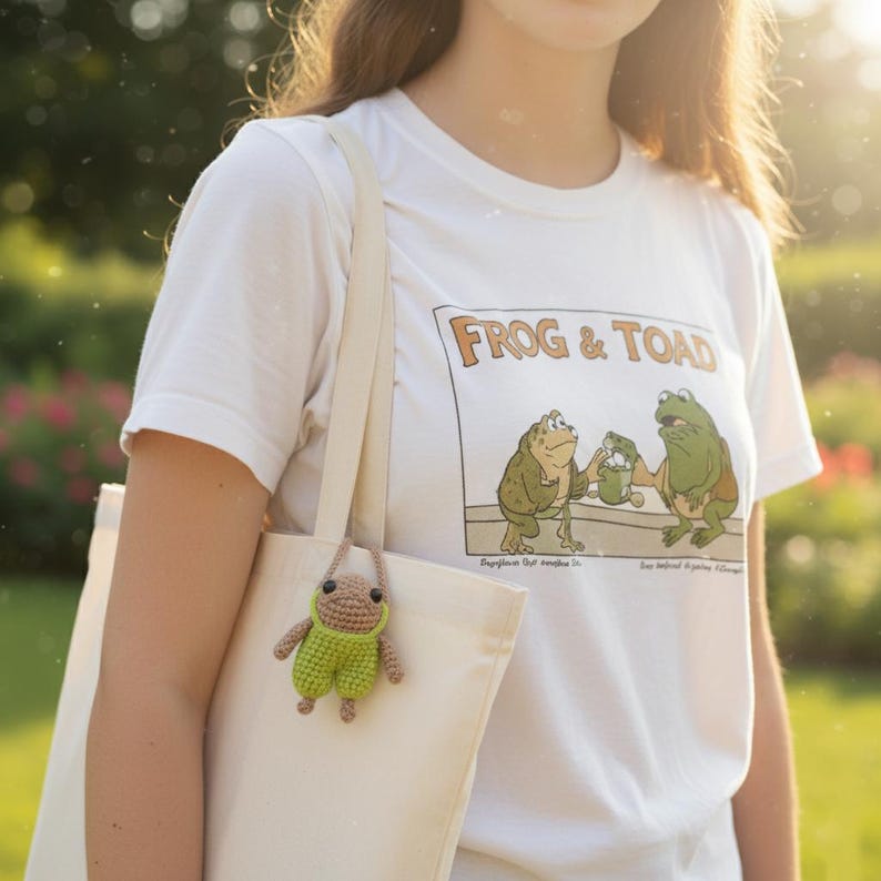 May include: A white t-shirt with a "Frog & Toad" graphic, paired with a cream-colored tote bag. A small, crocheted frog charm in green and brown hangs from the bag. The image is set outdoors with soft, natural lighting.