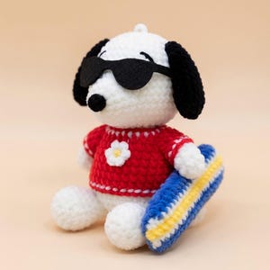 May include: A handmade crochet Snoopy doll wearing black sunglasses and a red sweater with a white flower. The doll is holding a blue and yellow surfboard. The Snoopy doll is white with black ears.
