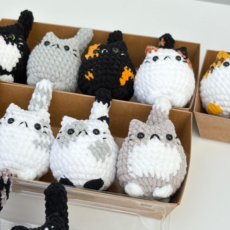 Puede incluir: Una colecci&oacute;n de figuras de gato de ganchillo hechas a mano en varios colores, incluyendo negro, gris y blanco, con acentos naranjas y amarillos. Los gatos se exhiben en cajas de cart&oacute;n individuales, mostrando sus dise&ntilde;os y texturas &uacute;nicos.